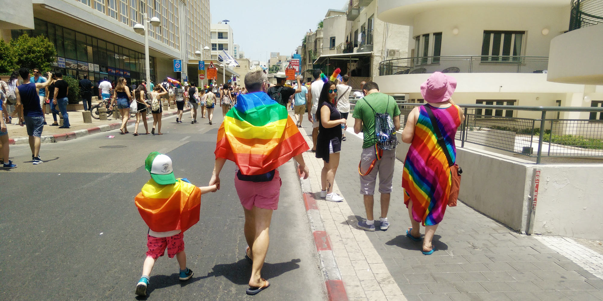 Tel Aviv Pride, father walks hand in hand with his child, both wearing rainbow flags, and a woman in a rainbow dress